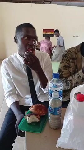 The video shows a group of people in what appears to be a large cafeteria or dining hall.  The room has a corrugated metal roof and a tiled floor. People are sitting at tables, eating and using their phones. Some have plastic bags with food. There are bottles of water on the tables. One man in a blue shirt and a patterned hat stands near the table and smiles. There are other people in the background.