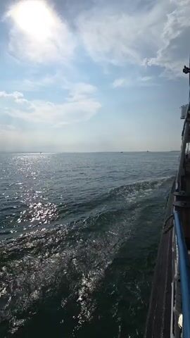The video shows a view from a boat sailing on the sea under a blue sky with scattered clouds. The sun is shining, casting reflections on the water. The camera is positioned to show the side of the boat and the wake it leaves behind. The water is a deep blue-green color. In the distance, there is a faint landmass or horizon line. There are no people visible in the frame.