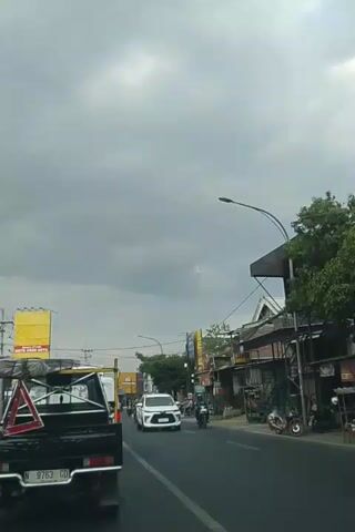 The video shows a view from a vehicle moving along a street. The sky is overcast and cloudy. On the right side of the street, there are buildings with shops and trees. There are several vehicles on the road, including a white car, a black pickup truck, and multiple motorcycles. People are visible riding on the motorcycles. The traffic appears to be moderate. The overall environment suggests a typical urban or suburban street scene.