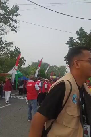 A group of people wearing red vests and white shirts are gathered on a street, some are facing a motorcycle and what appears to be a stall or tent decorated with Indonesian flags. The flags are red and white and are displayed on tall poles. A motorcycle is visible on the left side of the frame. Several people are standing around, some are talking, and a few are observing. The environment appears to be outdoors with trees and overhead wires. The sky is overcast.