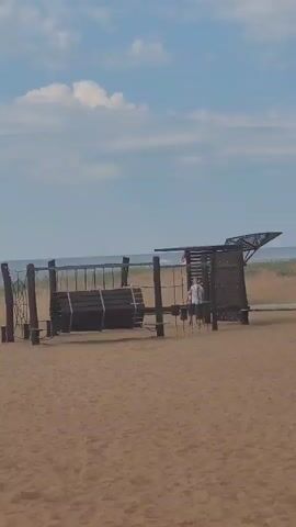 The video pans across a sandy beach with a playground structure. The playground features swings, a rope net, and a cylindrical object. Several people are visible on the beach and near the playground. In the background, the ocean stretches to the horizon under a cloudy blue sky. The camera movement is steady.
