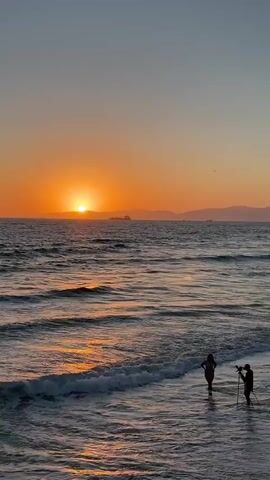 The video shows a sunset over the ocean. The sun is a bright orange circle low on the horizon, casting a warm glow on the water. The waves are gentle, with white foam cresting as they approach the shore. In the foreground, two people are standing in the shallow water. One appears to be a woman in a bikini, and the other is a person holding a tripod with a camera. They are facing the ocean, presumably watching the sunset or taking photos. In the distance, there are faint outlines of land and some ships on the water. The sky is a gradient of soft orange and blue, indicating the end of the day.