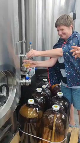 A woman is pouring a golden liquid from a large stainless steel tank into a glass. She is wearing a starry cape and shorts. Several large plastic jugs with white caps are lined up in front of the tank. Another person's hand is visible reaching towards the glass. The environment appears to be a brewery or similar facility, with another large metal tank visible in the background.