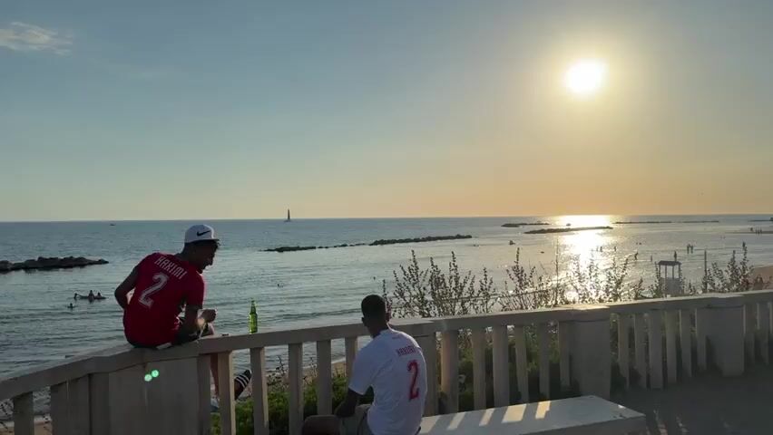 The video shows a scenic view of a beach during sunset. Two individuals are seated on a stone ledge in the foreground, facing the sea. One person, wearing a red shirt with the number 2 and the name HAKIMI on the back, is sitting on the edge of the ledge with their legs dangling. The other person, wearing a white shirt with the number 2 and the name HAKIMI on the back, is seated on a bench behind the ledge, also facing the sea. A bottle is visible on the ledge near the person in the red shirt. Beyond the ledge, the beach is populated with several people enjoying the sunset. Beach umbrellas and a lifeguard station are visible on the sand. The sun is setting over the calm sea, casting a warm glow and reflecting on the water. A sailboat is visible in the distance on the left side of the frame. The sky is clear and blue, transitioning to a lighter hue near the horizon. The environment appears to be a coastal resort or a public beach area.