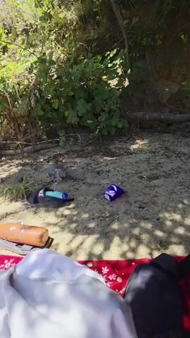 The video shows a gray bird foraging on the ground. A pair of black sandals is visible to the left of the bird, with a blue object next to them. In the center of the frame, a purple and blue foil wrapper lies on the ground. To the left of the wrapper, an orange water bottle is visible. In the foreground, a red blanket is spread out, with a white cloth and dark clothing on it. The background consists of dense green foliage and a dirt embankment. The scene appears to be outdoors in a natural setting.