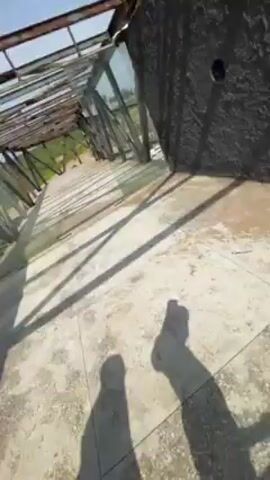 The video shows a person standing barefoot on a concrete platform, holding a dark, jagged rock in their hand. The background reveals a sandy beach with waves crashing ashore, and a small, dilapidated wooden structure is visible on the beach. The rock is held up against this backdrop.