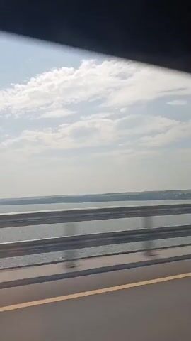 A video shows a view from a moving vehicle, likely a car, crossing a bridge over a body of water. The perspective is from inside the vehicle, looking out the side window. The scene is dominated by the water, the sky, and the bridge's railing. The water appears calm, with gentle ripples reflecting the sunlight. In the distance, the far shore is visible as a low, dark silhouette against the horizon, suggesting a landscape of hills or gentle slopes. The sky is a clear, pale blue, with a few scattered, wispy white clouds. The bridge railing, made of metal with horizontal bars, runs across the lower half of the frame, creating a sense of depth and movement. A yellow stripe is visible on the road surface below the railing. The overall impression is one of a journey on a bright, clear day, with the vastness of the water and sky providing a sense of open space. No people are visible within the frame. The video appears to be a short clip, possibly a segment from a longer travelogue or dashcam footage.