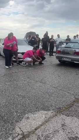 The video shows several people gathered around a race track. Two men wearing pink shirts and shorts are squatting down to inspect the ground in front of a silver BMW. The BMW has a license plate that reads "PB 7103HE." Behind the BMW is a silver Subaru with a license plate that reads "M 1179 BP." Several people are standing behind the men in pink shirts. Most of them are wearing black shirts and shorts or pants. Many of them are holding up their phones and recording. A third car, a green BMW, can be seen entering the frame from the left side of the screen. The sky is cloudy and the ground is paved. The audio is loud and sounds like the engines of race cars.