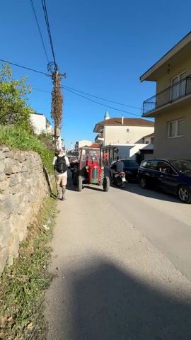 A man with a backpack is walking down a narrow street, away from the camera's point of view. On the same street, a red tractor, pulling a trailer covered with a green tarp, drives toward the camera. The street is lined with buildings on both sides, and several cars are parked on the right side. The sky is clear and blue. There are speed bumps on the street. A vine is wrapped around a utility pole.