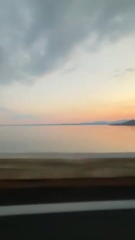 A video taken from a moving vehicle shows a body of water and a sky with clouds. The water is calm and reflects the colors of the sunset, which include pinks, oranges, and purples. The sky is mostly gray with some pink and orange hues near the horizon. The vehicle appears to be traveling along a road next to the water. There are some trees visible on the side of the road, and the movement of the vehicle creates a blurred effect in the video. The overall mood of the video is peaceful and serene.
