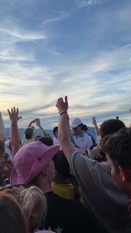 The video shows a crowd of people gathered together outdoors during what appears to be sunset. The sky is filled with dramatic clouds, and the sun is setting behind a mountainous landscape, casting a warm glow over the scene. Many people in the crowd are holding up their hands, some are holding phones or cameras, possibly to take pictures or videos of the sunset or the event. A few individuals are wearing hats. The general atmosphere seems to be festive and celebratory. The camera angle is from within the crowd, looking upwards towards the sky and the distant scenery.