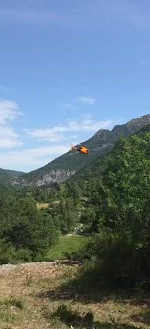 A small orange helicopter flies through a clear blue sky over a lush green mountainous landscape. The helicopter has a rotor and is suspended by a long cable, from which a rectangular object is hanging. The object appears to be a cargo container or basket. The helicopter is moving from left to right across the frame. The terrain below is heavily forested with steep, rocky mountains in the background. The shot is taken from a low angle, looking up at the helicopter and the sky.
