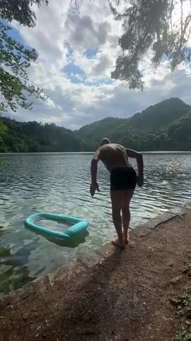 The video shows a person in black swim trunks diving into a lake. The person is seen bending over the edge of the bank and then jumping into the water. An inflatable pool float is in the water near where the person jumps. The lake is surrounded by green, tree-covered hills and the sky is cloudy. The video lasts for 4 seconds.