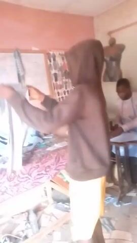 A young man wearing a brown hoodie and yellow shorts stands in a room, holding up a white piece of fabric. He is facing the camera, looking at the fabric. In the background, another man is sitting at a sewing machine. There is a bed covered with fabric on the left, and a mannequin is visible in the corner. The room appears to be a tailor shop.