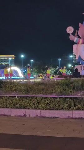 A group of people, appearing to be children and adults, are participating in an outdoor activity at night. Most of the individuals are wearing brightly colored athletic attire, predominantly yellow tops and pink shorts. They are arranged in a semi-circle, facing a tall, illuminated structure that emits a bright green light. The individuals are engaged in synchronized movements, with many of them raising their arms and stretching. The environment is an urban park or public space, indicated by the paved walkways, greenery, streetlights, and buildings in the background. One person is seated on a bench to the right of the frame, facing away from the camera. A bicycle is parked to the right of the performing group. In the background, a large, stylized cartoon-like figure, possibly a pig, is visible, holding a large object resembling a fan or shield. The overall atmosphere is festive and energetic, with the bright lights contributing to the visual spectacle.