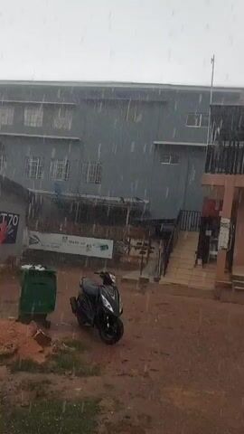 The video captures a rainy outdoor scene with a multi-story building in the background. In the foreground, a black scooter is parked on a muddy ground, next to a green garbage bin. The building appears to be an institutional or commercial structure with multiple windows. The rain is visible as streaks across the frame, indicating moderate to heavy precipitation. The overall atmosphere is damp and overcast, typical of rainy weather.