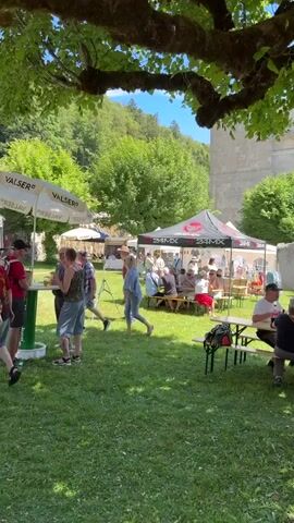 The video shows an outdoor gathering on a sunny day under a large tree with green leaves. Several people are present, mingling and standing around tables and tents. To the left, under a white umbrella with the "VALSER" logo, a group of people are gathered around a green table. In the background, there are tents, including one with a "24MX" logo, and people are seated at picnic tables. A young child is sitting on a tricycle in the foreground. The environment appears to be a grassy area with trees and a stone building in the background.