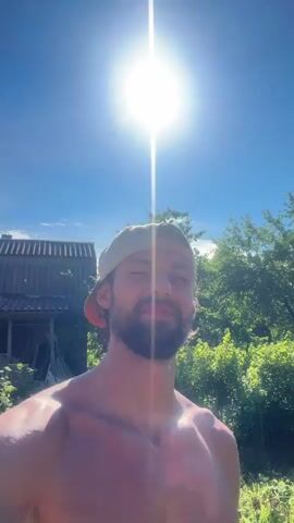 The media is a selfie-style video of a shirtless man with a beard, wearing a baseball cap. He is outdoors in a sunny environment. The background includes lush green foliage and a rustic, unfinished building, possibly a shed or small house, constructed of cinder blocks and wood. The sun is bright, suggesting it is daytime, likely late morning or afternoon, and the weather appears warm and clear. The man has a relaxed and contented expression, with a slight smile. He is in Kuti, Russia, and the scene conveys a feeling of being in a rural or semi-rural setting. There is no visible text in the video.