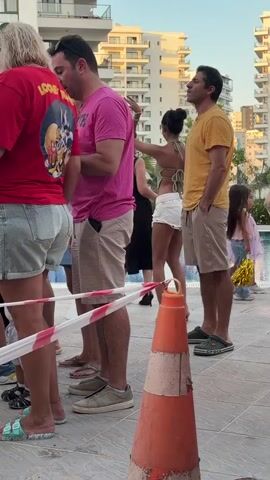 Four people are standing in a line. A woman wearing a red shirt with "Looney Tunes" printed on it is on the left. Next to her is a man in a pink t-shirt and khaki shorts. Behind him, a woman in a white top and shorts is talking to a man in a yellow t-shirt and khaki shorts. A traffic cone is in the foreground. There is a swimming pool in the background.