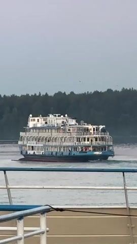 A large cruise ship with multiple decks moves across a wide body of water. The ship is white and blue with red trim along its waterline. Dense green trees line the distant shore. The sky is overcast. The video appears to be filmed from another boat, as the railing and deck of a second vessel are visible in the foreground.