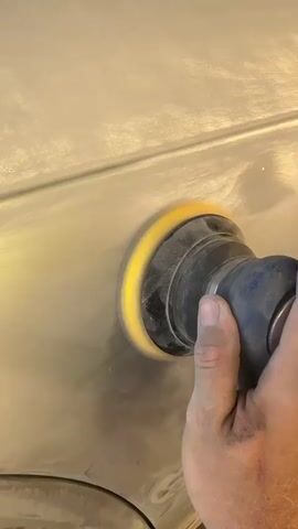A close-up shot shows a hand holding a sander with a yellow sanding pad. The sander is being used to sand a light-colored surface, likely a car body panel, which has a faint horizontal line running across it. Dust is being generated by the sanding action. The lighting is bright, highlighting the texture of the surface and the motion of the sander.