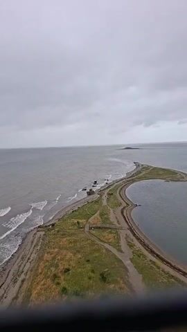 The video shows an aerial view of a coastline with a narrow strip of land extending into the ocean. The water is grey and choppy, with waves crashing onto the shore. There are some scattered rocks and a few concrete structures along the coastline. The land is covered in dry grass and has several dirt paths winding through it. In the distance, a small island is visible. The sky is overcast and the overall atmosphere is muted. There are a few people walking along the paths.
