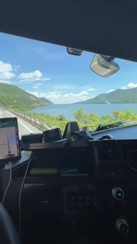 The video shows the view from inside a truck cab, looking forward through the windshield. A large body of water, likely a lake or bay, is visible on the right, bordered by lush green, mountainous terrain. The sky is blue with scattered white clouds. A paved road runs along the left side of the water, and there are other vehicles visible on the road, including a bus and what appear to be other trucks. The truck cab interior shows a dashboard with various instruments and devices, including a smartphone mounted on the left, displaying a navigation map. The driver's steering wheel is partially visible on the left. The scene suggests a scenic drive along a coastal or lakeside highway on a clear, sunny day.