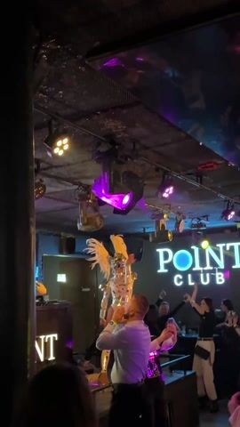 In a dark nightclub setting in Bydgoszcz, Poland, a performer in an elaborate, futuristic white and silver costume with large white feathered wings and a helmet mask dances on stage under bright stage lights. The costume also features gold accents and appears to be a full-body suit with reflective elements. Several audience members are visible, with some filming the performance on their phones. One man in a white shirt stands close to the stage, watching intently. Behind the performer, a large illuminated sign reads "POINT CLUB," with part of another "T" sign visible to the left. The stage is lit with various colorful lights, predominantly green and purple, creating a vibrant atmosphere for the live show.