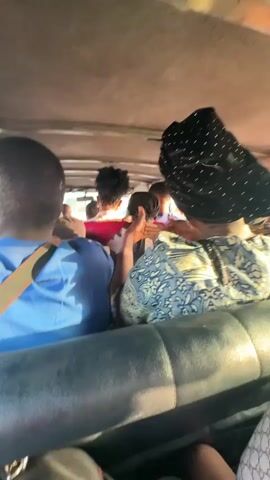 The video depicts the interior of a moving vehicle, possibly a minibus or van, filled with passengers. The viewpoint is from the back, looking forward towards the front. Several people are visible, including individuals in a blue shirt with a seatbelt, a woman in a blue and white patterned top wearing a black head wrap with small white dots, and others. The woman in the patterned shirt appears to be pointing or gesturing towards the front of the vehicle. The ceiling of the vehicle is low and light-colored. The seats are dark, possibly leather or vinyl, showing signs of wear. The vehicle appears to be in motion. Loud audio is present.