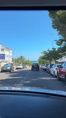 The video is a first-person perspective shot from inside a car, looking out through the windshield. The car is on a street with two lanes of traffic, lined with parked cars on both sides. The street is paved and has white dashed lines marking the lanes. There are trees and buildings along the sides of the street, and a clear blue sky above. In the distance, a body of water, likely the ocean, is visible. The sun is shining, casting shadows on the road. The camera is relatively still, indicating the car is moving at a steady pace. Several cars are visible on the road, with one black SUV directly in front of the camera car. The environment appears to be a coastal town or city.