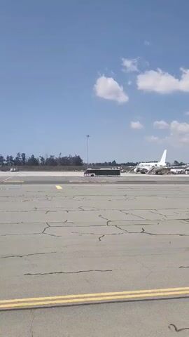 The video shows an airport tarmac on a sunny day with a blue sky and scattered white clouds. In the foreground, the cracked asphalt of the runway is visible with yellow lines marking the edge. In the mid-ground, a black bus is driving across the tarmac. Further in the background, a white airplane is parked at a gate, connected to a jet bridge. There are also other airport vehicles and infrastructure visible. Trees line the horizon behind the runway.