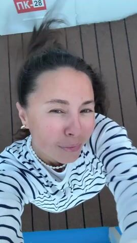 A woman with long brown hair is shown from a low angle, looking upwards. She is wearing a striped long-sleeved shirt and light-colored pants. Her hair is being blown upwards by the wind, obscuring part of her face in some frames. She smiles and makes expressions at the camera. The background shifts between a wooden deck with a red and white sign that reads PK-28 and a bright blue sky. The camera movement is dynamic and jerky, suggesting it is being held by someone in motion.