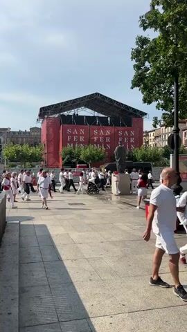 The media file depicts an outdoor public square filled with people. The central focus is a large stage draped in red fabric, with large white lettering that reads "SAN FER MIN" repeated multiple times. In front of the stage, a statue of a person stands on a pedestal.

The crowd is predominantly dressed in white clothing with red accents, such as scarves or sashes, suggesting a festive or ceremonial event. Many individuals are walking, some in groups, while others appear to be engaged in casual conversation or movement. A few people are holding bags or pushing strollers. Two red trash receptacles are visible in the foreground. The ground is paved with large, light-colored tiles.

The scene appears to be set during the daytime, under a bright, partly cloudy sky. The presence of trees with green foliage suggests a warm-weather season. The architecture of the surrounding buildings, with their balconies and tiled roofs, hints at a European setting, specifically Pamplona, Spain, as indicated by the "SAN FER MIN" text, which is likely a reference to the San Fermín festival. The overall atmosphere seems lively and celebratory, with a sense of community gathering.