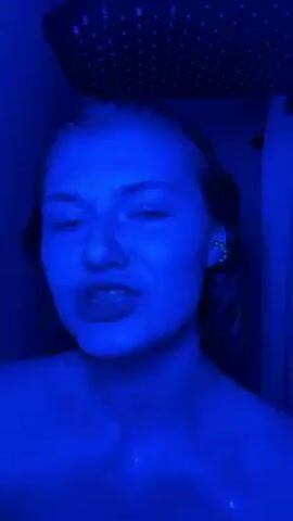 A young fair-skinned woman with wet hair and light-colored eyes is featured in a close-up, chest-up shot under blue lighting, likely in a shower. A chrome shower head with small holes is visible above her, while a shower curtain hangs to her right. She touches her face and mouth with her finger while looking directly at the camera and singing. The environment is bathed in a saturated blue light. She is wearing earrings.
