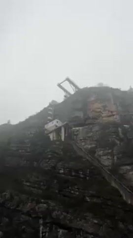 The camera pans up along a rocky cliff face, revealing a funicular structure and a dam wall. The funicular includes a steep railway track climbing the mountain, a small white building, and an upper station structure. The dam wall is large and curved, standing out against the foggy sky.  The overall environment is rugged and dominated by the natural rocky landscape.