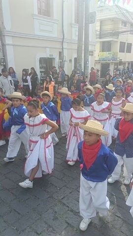 Children are marching in a parade down a cobblestone street. They are wearing traditional costumes, including white dresses with red trim for the girls, and blue or white shirts with red or blue bandanas for the boys. Many of them are wearing straw hats. There are people watching the parade from the sidewalks on either side of the street. The buildings are mostly light-colored. Banners with a series of colorful triangles are hung between some of the buildings. The weather appears sunny.
