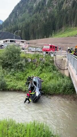 A dark-colored car is partially submerged in a shallow river, tilted on its side. Several firefighters, wearing helmets and protective gear, are working to stabilize and potentially move the vehicle. Some firefighters are in the water near the car, while others are on the bank. A red fire truck is visible in the background, along with a bridge and a wooded hillside. The scene appears to be a rescue operation.