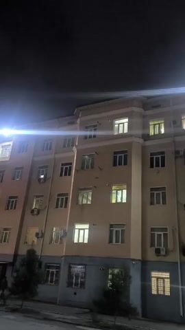 The video shows a five-story residential building at night. The building is beige with a gray base. Windows are visible on each floor, some with lights on inside. Several air conditioning units are attached to the building.  Two small trees are planted in front of the building. The sky is dark.  There are street lights in the background. The video appears to be taken from across the street.
