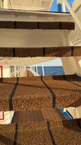The video shows a close-up view of textured, brown stairs with black stripes. The stairs are part of a larger structure, possibly a boat or a pier, as evidenced by the white railings and the view of the ocean and sky beyond. The camera pans upward, revealing the vast expanse of the blue ocean under a clear sky. In the distance, a lighthouse stands on a rocky island, silhouetted against the horizon. The water appears calm, with gentle waves. The lighting suggests it is daytime, possibly late afternoon or early morning given the soft light. There are no people visible in the frame. The overall scene conveys a sense of tranquility and the immensity of nature.