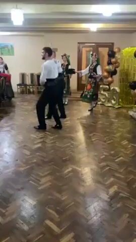 In a medium shot, a group of dancers, two women and two men, are performing a traditional folk dance in a spacious room. The dancers are wearing traditional costumes, with the women in colorful floral skirts, white blouses, and headscarves, and the men in black pants, white shirts, and black vests. The room has a parquet floor and high ceilings, with a chandelier visible in the background. There are chairs and tables along the walls, and a balloon display with the number "50" is visible in the background, suggesting a birthday celebration. The camera is stationary, capturing the dancers as they move gracefully across the floor. The overall tone of the video is festive and celebratory, highlighting the beauty and energy of the traditional dance.