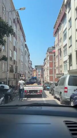 A white Kia Motors truck is driving down a narrow street. The truck is loaded with dirt or sand and is being driven by a person who is not visible. The truck is being followed by a silver van and other vehicles. The street is lined with apartment buildings and shops. The sky is clear and blue.