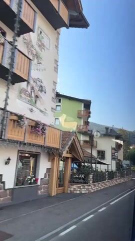 A video captured from inside a vehicle shows a street scene in a European town. On the left side of the road, there are buildings with traditional architecture. The first building is light-colored with a pitched roof and wooden accents. Next to it is a green building with balconies, followed by a cream-colored building with several windows and balconies. A sign for a pizzeria is visible on this building. Further down the street, there are more buildings, some with intricate architectural details and decorative elements. There is a sidewalk and a road with white lane markings. Trees and green spaces are present alongside the road. A black car with a roof rack is parked in front of one of the buildings. The sky is clear and blue, indicating daytime. The video appears to be filmed as the vehicle moves along the street, offering a continuous view of the town's facade.