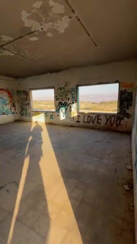 The video shows the interior of a room or building with graffiti covering the walls and floor. Sunlight streams in through two windows, casting a long shadow across the room. A young woman wearing a white top and a yellow skirt walks away from the camera towards a doorway. Another person, a man in a blue t-shirt, is seen walking ahead of her through the doorway into another area of the building. The environment appears to be abandoned or derelict, with peeling paint and a generally unkempt appearance.