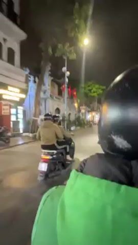 The video shows a night scene from the perspective of someone riding on the back of a scooter. The person in front is wearing a black helmet and the videographer is wearing a bright green jacket. They are driving down a narrow street lit by streetlights. There are buildings lining the street, some with shops and lights visible.  Several people are walking on the sidewalk. There is at least one Vietnamese flag visible. Other scooters are parked on the side of the road and driving nearby.