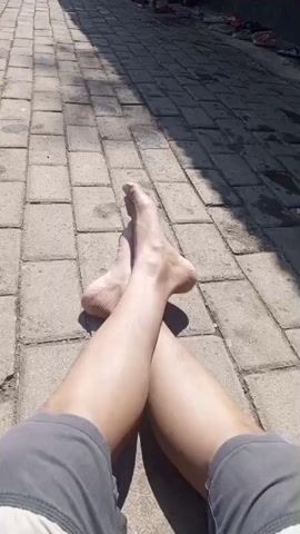 The video begins with a close-up shot of a person's legs and feet, bare and crossed, resting on a paved walkway composed of rectangular bricks. Sunlight casts shadows on the bricks, indicating it is daytime. The camera then pans to the left, revealing a water park scene. Children and adults are visible in a shallow pool with various water play structures, including arches with sprinklers and dolphin-shaped fountains. Lush green trees and plants surround the pool area, and a paved pathway continues along the edge of the park. The sky is partly cloudy, and the overall atmosphere appears to be one of leisure and recreation.