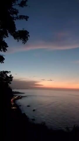 The video shows a sunset over the ocean. The sky is a deep blue at the top, transitioning to lighter shades of blue and pink near the horizon. The ocean is calm and reflects the colors of the sky. In the foreground, there are silhouetted trees and a coastline with lights from a town. The camera is stationary and the video is edited with several short clips that show the same scene from slightly different angles or with subtle shifts in perspective.