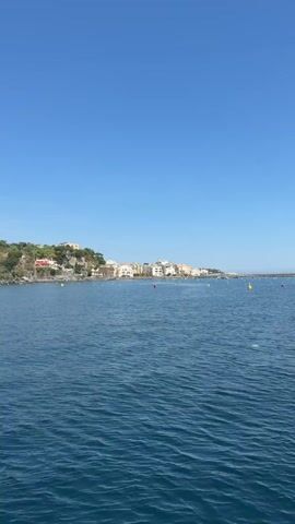 The camera captures a daytime view of a body of water with a coastal town in the distance. The water is a deep blue with gentle ripples across its surface. Several buoys, colored yellow and red, are visible in the water, indicating potential navigational markers. On the left side of the frame, a coastline is visible with a cluster of buildings, primarily white with some orange roofs, nestled at the base of a green, tree-covered cliff. To the right, another land mass rises from the water, also covered in trees and topped with buildings. The sky above is a clear, cloudless blue. The scene exudes a tranquil, seaside atmosphere.