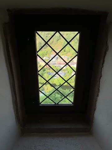 This video shows a view through a diamond-paned window looking out onto a village nestled in a valley. The window frame is dark wood, and the interior wall around the window is a rough, light-colored material, possibly stone. Outside, the village has houses with red-tiled roofs, surrounded by lush green trees. A river or body of water is visible, reflecting the sky. The landscape appears to be hilly, with more greenery and structures visible on the slopes. The diamond panes of the window are made of glass, with thin black metal framing. The scene is well-lit by natural light coming from outside.
