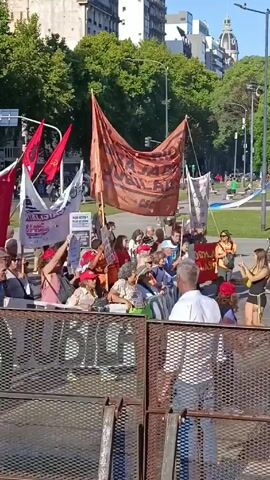 The video shows a crowd of people marching down a city street, presumably a protest or demonstration. They are holding banners and flags, some of which are red. The crowd is mostly composed of older adults. They are moving behind a temporary metal barrier. The street is lined with trees and buildings, and the sky is bright and sunny.