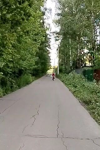 A young person is riding a bicycle down a paved road surrounded by trees. The person is wearing a red shirt and is carrying a plastic bag. The road is cracked and appears to be in disrepair. The trees on either side of the road are lush and green. The sky is overcast. The scene appears to be a rural or suburban area.